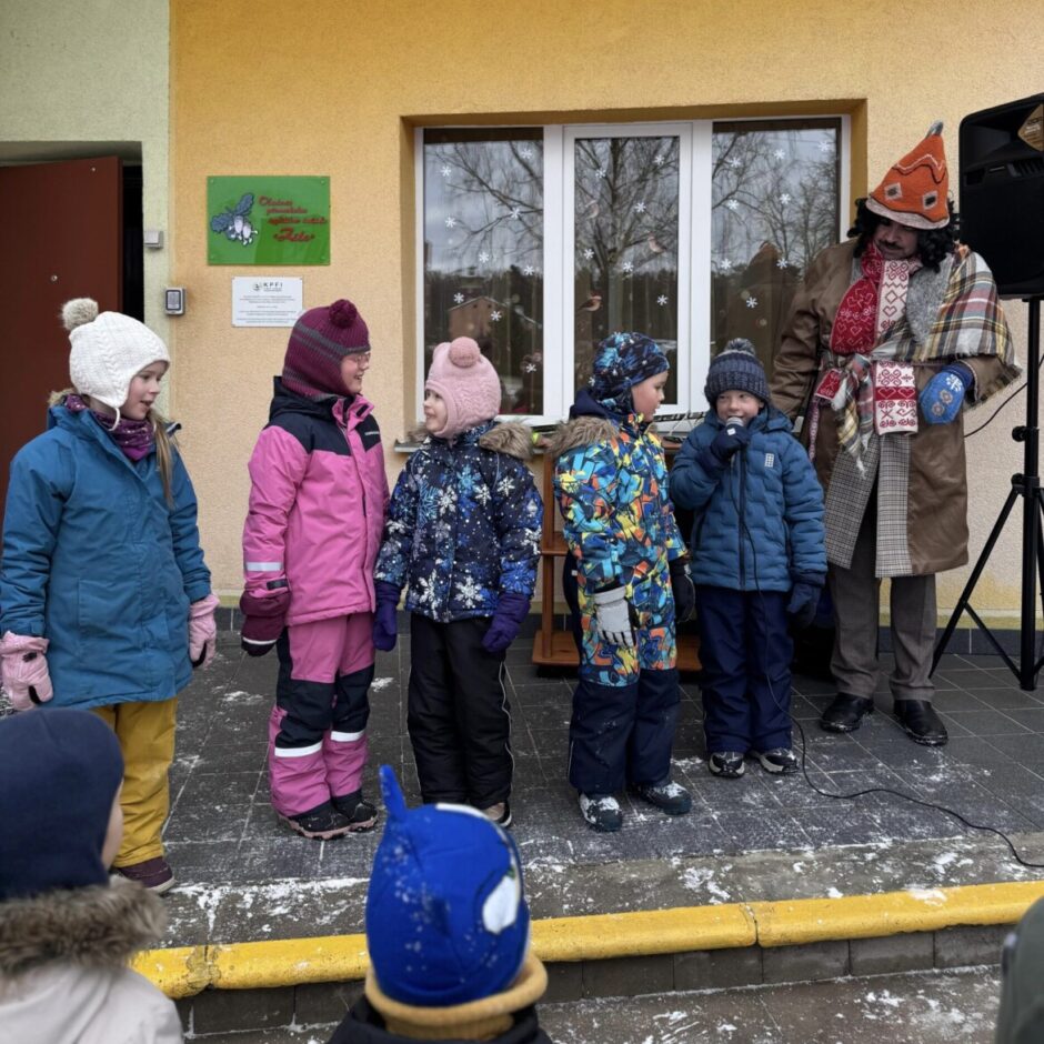 Mazs bērns zilā ziemas apģērbā un cepurē ar dinozaura radziņiem no muguras spēlējas sniegā, turot rokās krāsainu, taisnstūra rāmi ar punktiņu rakstu.