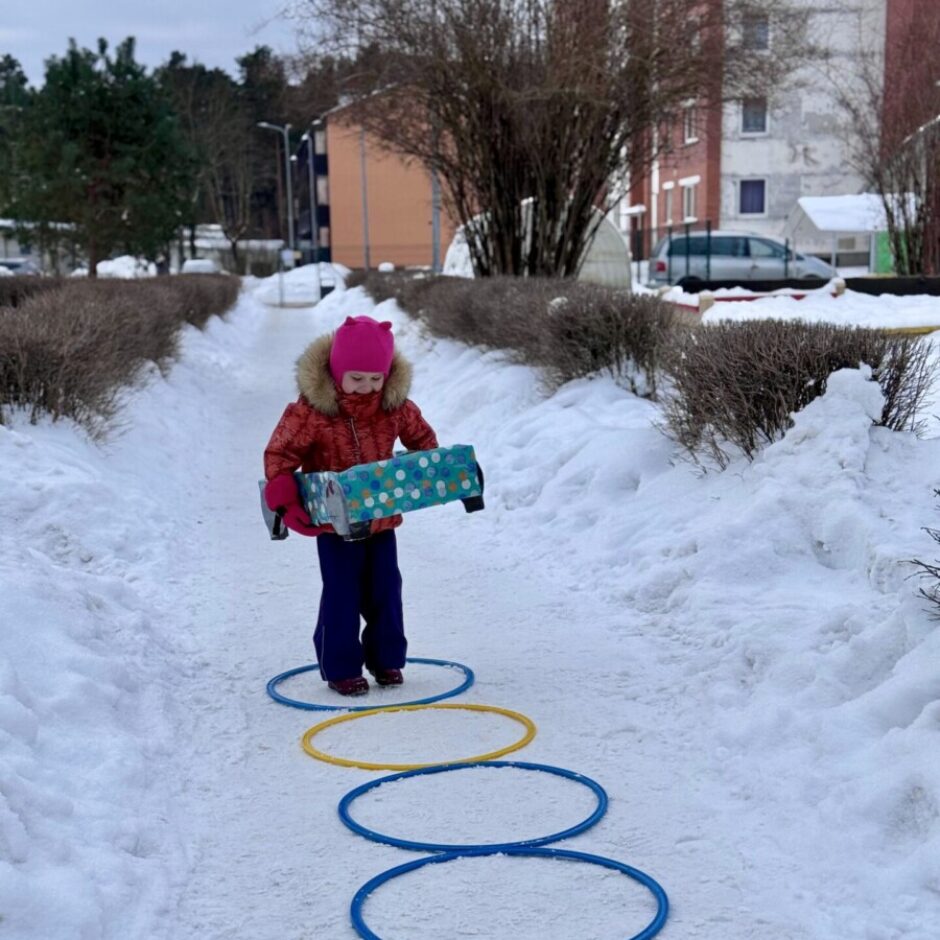 Maza meitene sarkanā jakā un rozā cepurē iet pa sniegotu taciņu, turot rokās iesaiņotu dāvanu. Uz taciņas ir izvietoti zili un dzelteni riņķi, veidojot celiņu. Apkārtne ir klāta ar sniegu, fonā redzami krūmi un ēkas.