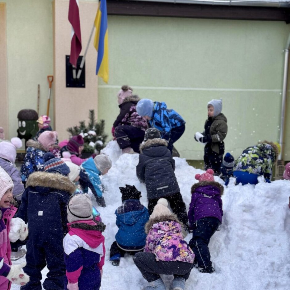 Bērni krāsainos ziemas apģērbos aktīvi spēlējas ar sniega kaudzi laukā pie gaiši zaļas ēkas, pie kuras plīvo Latvijas un Ukrainas karogi.