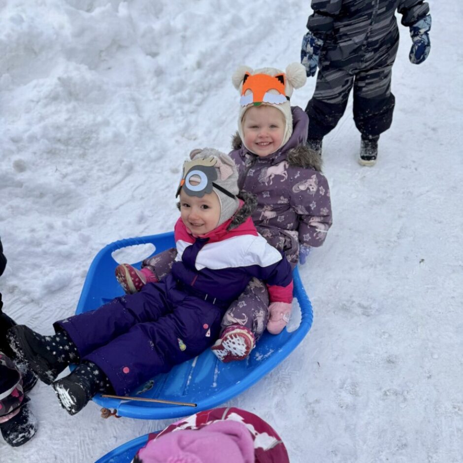 Divi mazi bērni smaidot sēž uz zila šļūcamā dēlīša sniegā. Viens tērpies lapsas cepurē, otrs — cepurē ar Miniona brilles motīvu. Aiz viņiem stāv cits bērns, un priekšplānā redzams vēl viens bērns uz cita dēlīša.