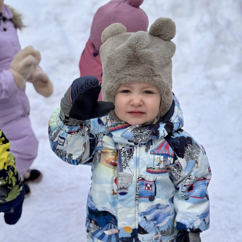 Mazs bērns ziemas apģērbā ar lāču ausu cepuri stāv sniegā, vicina roku un skatās kamerā. Fonā redzami citi bērni.
