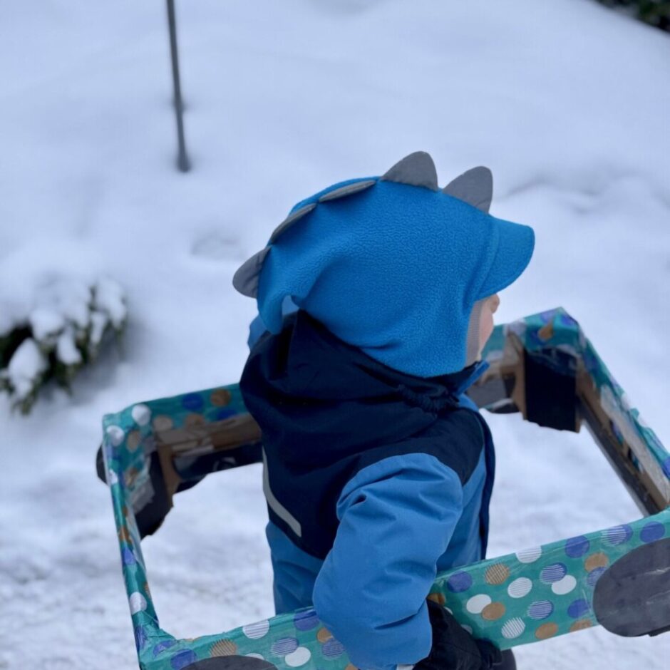 Mazs bērns, ģērbies zilā ziemas kombinezonā un cepurē ar dinozauru radziņiem, iet pa sniegu, turot rokās krāsainu, ar punktiņiem rotātu taisnstūra rāmi.