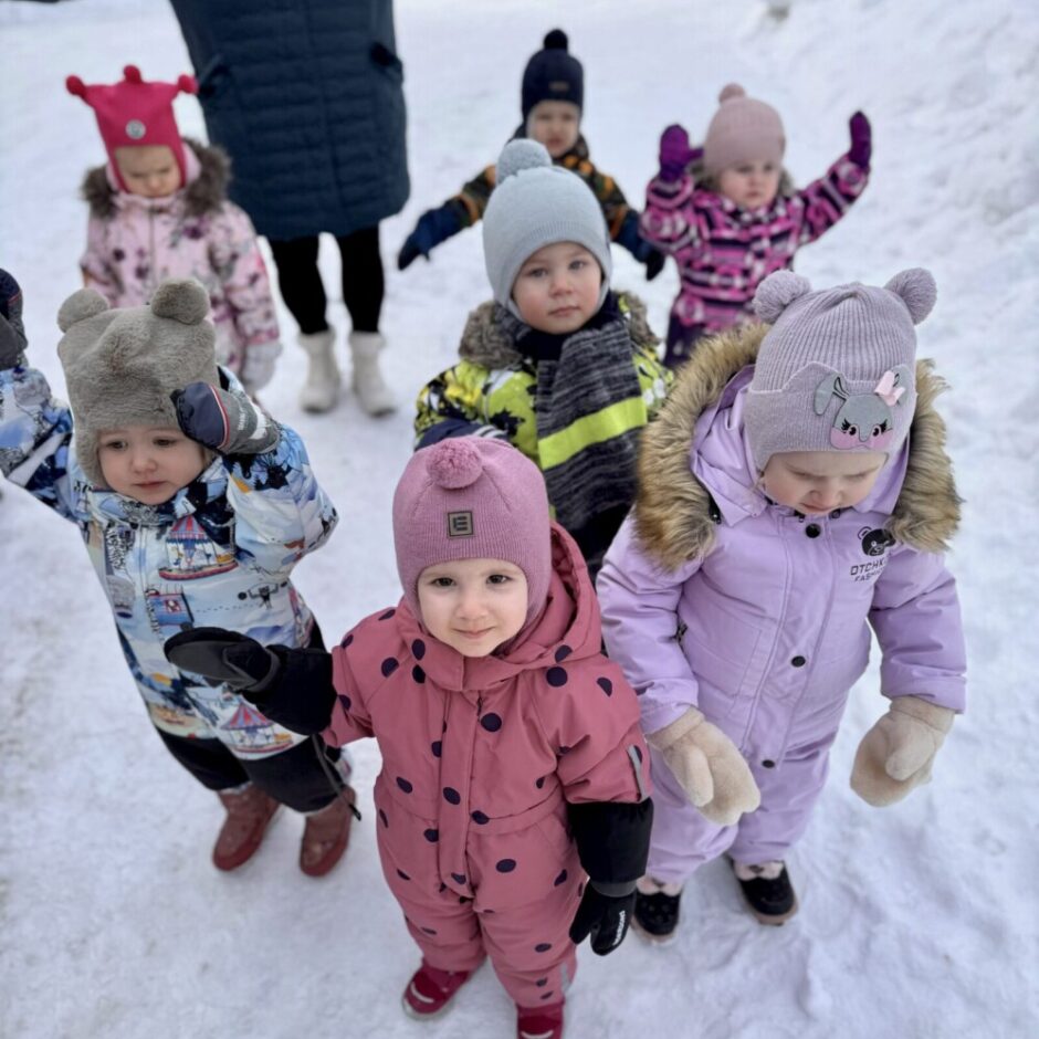 Septiņi mazuļi krāsainos ziemas kombinezonos un cepurēs stāv sniegotā āra vidē, fonā daļēji redzams pieaugušais. Priekšplānā esošais bērns rozā kombinezonā ar punktiņiem skatās kamerā.